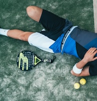 A person is lying on the ground, wearing athletic clothing that includes a blue and black top and white shorts. Nearby, there is a paddle racket and two yellow balls on a textured surface, which looks like a sports court. The person is wearing black sneakers and has a wristband on their arm.